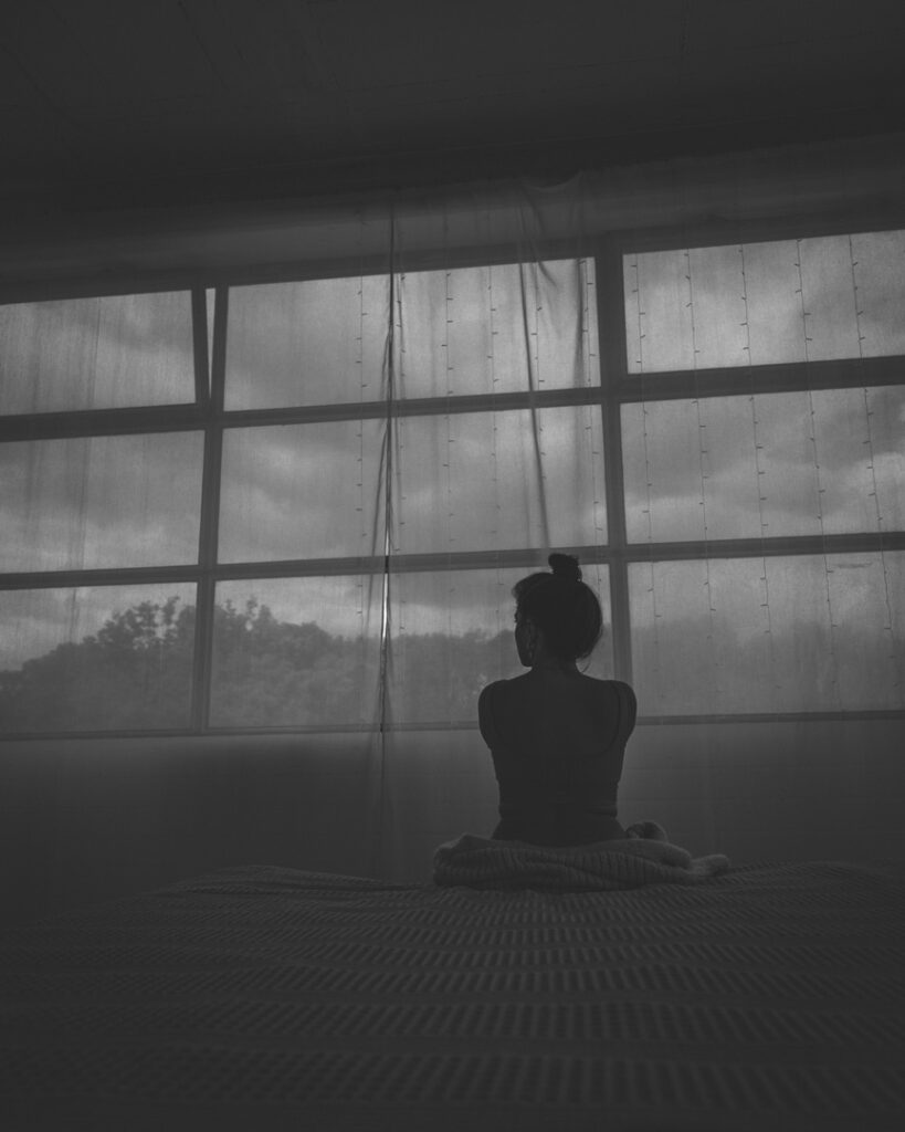 Frau sitzt auf Bett und wendet der Kamera den Ruecken zu